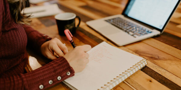 Girl sitting at a table in front of a laptop writing notes on creating powerful content in a notebook