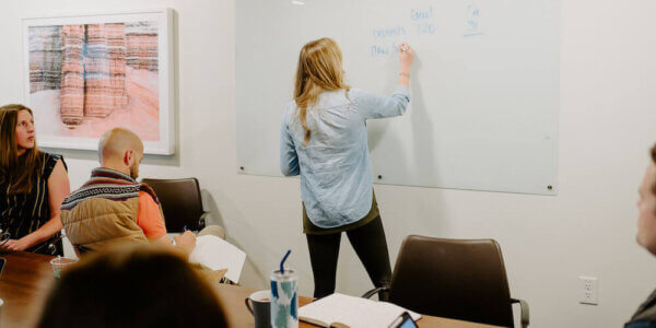 Kate writing on a whiteboard about delivering value with the team at a table behind her