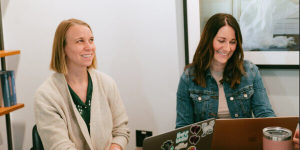 Two women smiling while sitting at a table with laptops in front of them