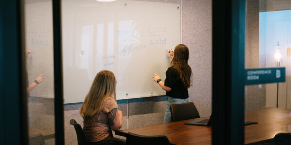 Looking into a work room with two girls looking at a whiteboard, one of them writing on the whiteboard about how to think about data while the other sits down looking