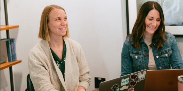 Two women smiling sitting at a table with laptops discussing why you shouldn't stop marketing