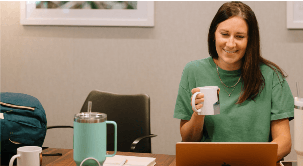 Woman sitting at table holding coffee cup typing on laptop about AI visibility